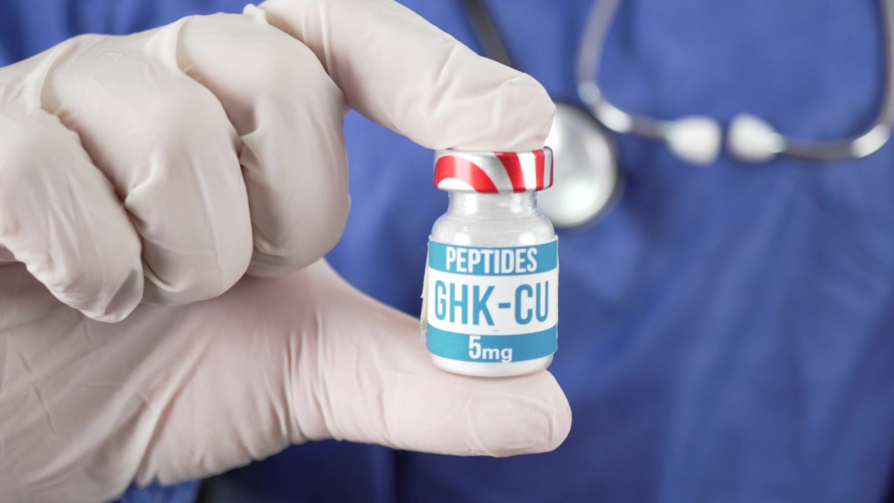 Dita guantate di lattice bianco tengono una bottiglia etichettata di peptide GHK-CU da 5 mg davanti a una persona che indossa uno stetoscopio