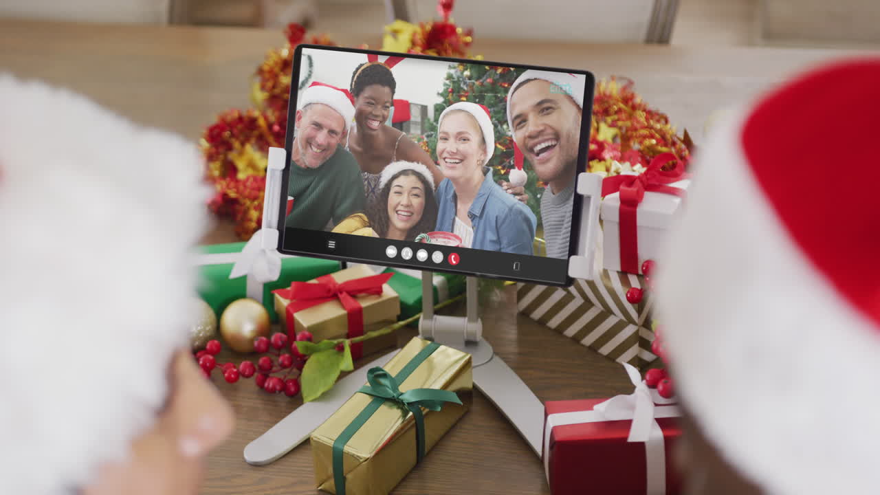 una pareja caucásica con sombreros de santa haciendo una llamada de video de tableta con amigos felices y diversos