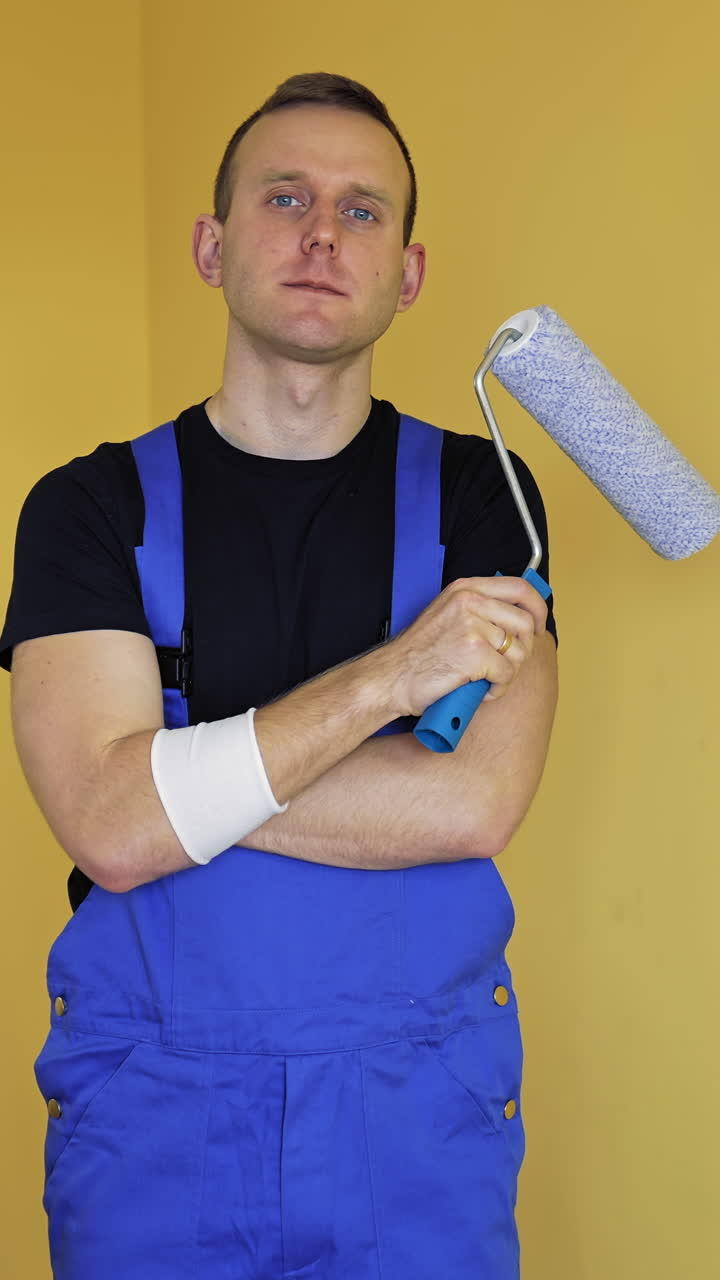 Portrait of a worker painter. Young man in blue overalls standing in the room and holding roller brush before doing a makeover in flat. Home improvement and renovation. Vertical video