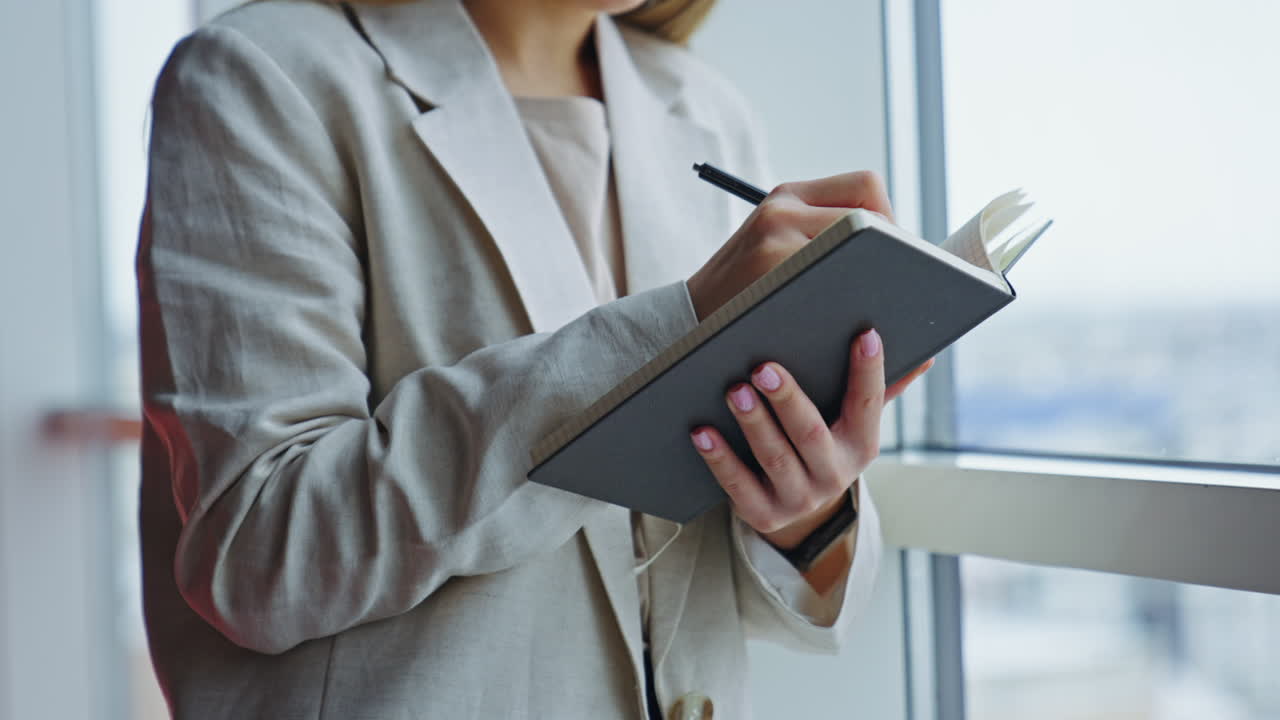 Lady in light jacket standing at the window and holding paper notebook. Woman making notes in her diary. Close up.