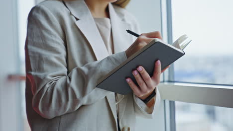 lady in light jacket standing at the window and holding paper notebook. woman making notes in her diary. close up.