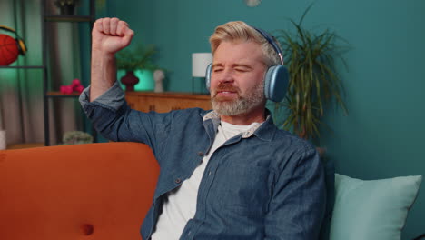 hombre maduro feliz con auriculares inalámbricos relajándose sentado en el sofá en casa escuchando su música favorita