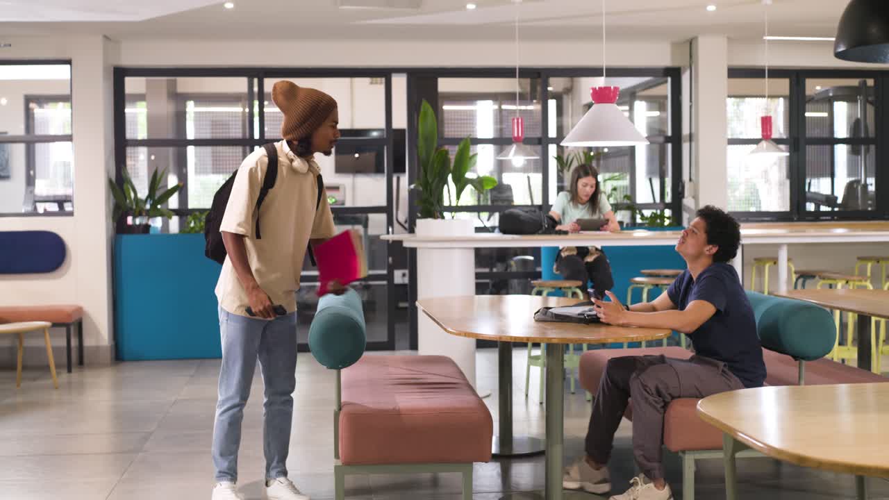 African American man entering campus lounge from left placing pink folder greeting peer thumbs-up