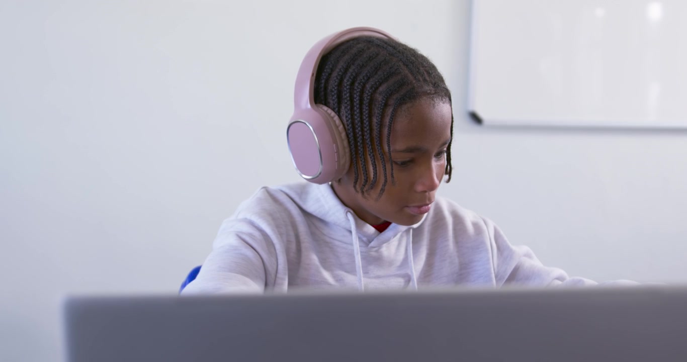 African American teenager focusing on laptop at study desk, lifting pink headset, adjusting, typing