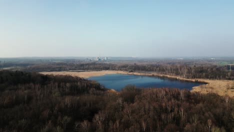 Serene-landscape-featuring-a-blue-lake-surrounded-by-brown-and-green-vegetation,-with-distant-buildings-under-a-clear-sky