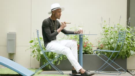 Wealthy-man-wearing-fedora-hat-and-gold-jewelry-sits-at-a-table-drinking-wine-holding-a-cigar-outside-near-green-plants