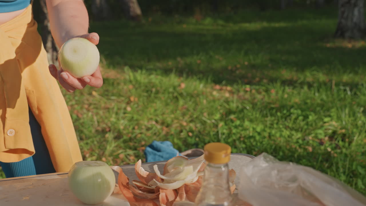Camping cooking woman slicing onion outdoors closeup hands peeling and chopping onion on picnic table with knife, bowl, jar and onion skins, warm sunlight over grassy campsite, yellow skirt and blue