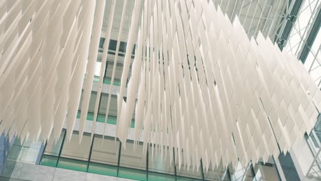 low angle shot of a white chandelier cascades hanging along high roof of a building with glass walls in the background