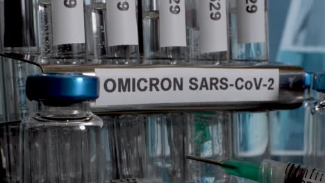 a close look on a  metal tube rack labeled omicron sars-cov-2. a metal tube rack contains glass test tubes named b.1.1.529. a few syringes lie in front of a rack. the background is blurry