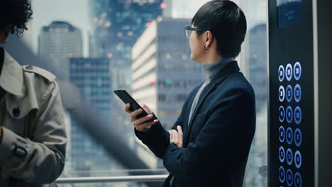 successful stylish japanese businessman riding glass elevator to office in modern business center. handsome man smile while using smartphone, write text message, check social media in a lift.