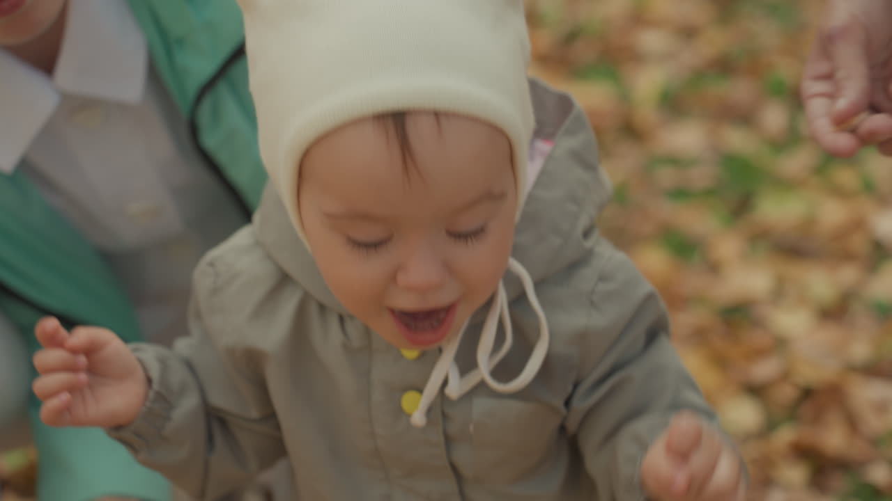 Baby reaches for parent hand autumn park, caregiver kneels guiding tiny steps through crisp brown leaves, knit hat and cozy jacket, closeup on shoes and curious fingers, tender bonding moment