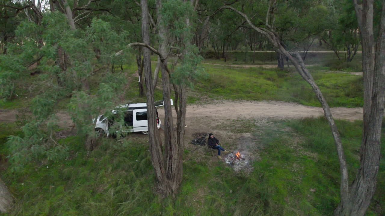 A lone camper warms himself beside a fire on a riverbank in the Australian bush.