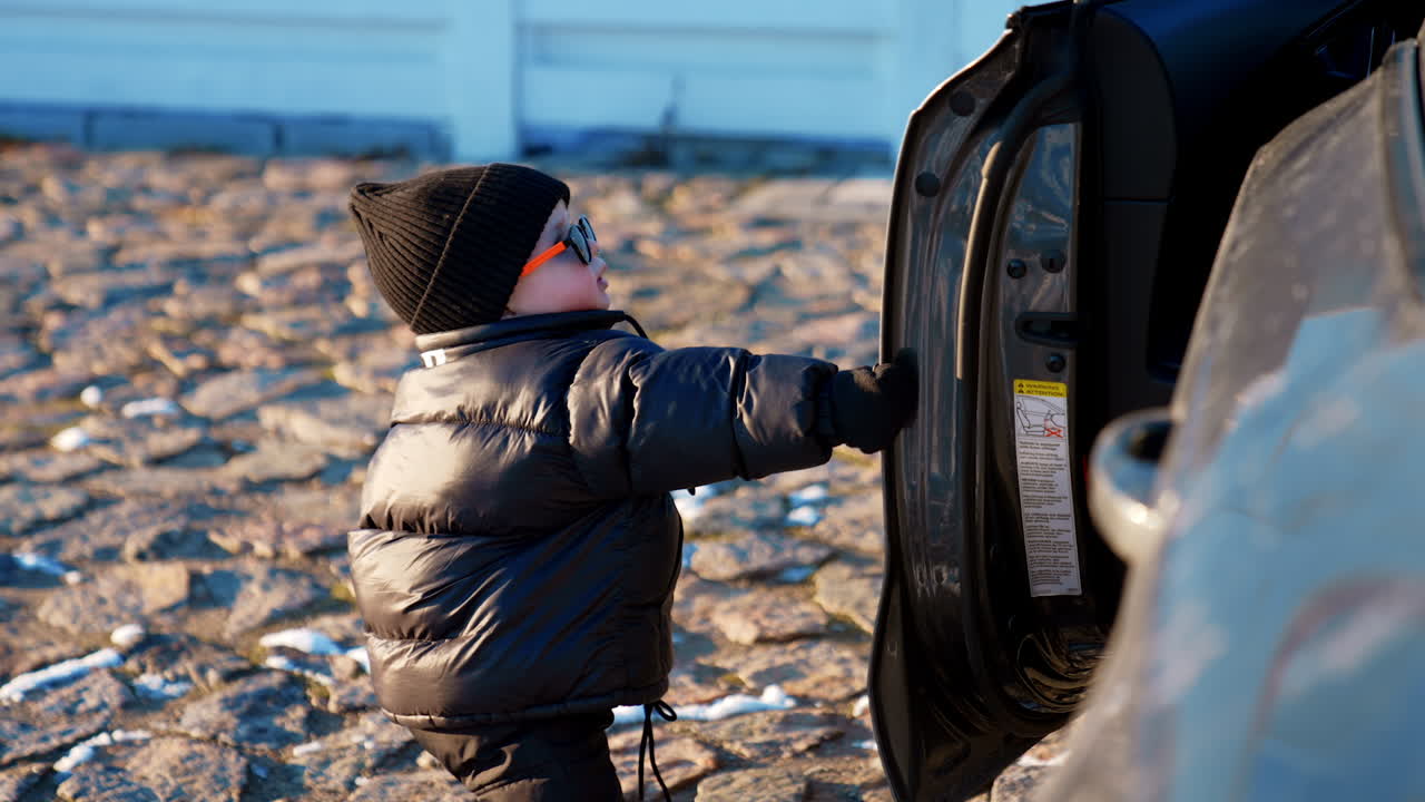 Lovely modern baby boy in sunglasses stands at the car. Toddler kid pulls the door open of the vehicle.