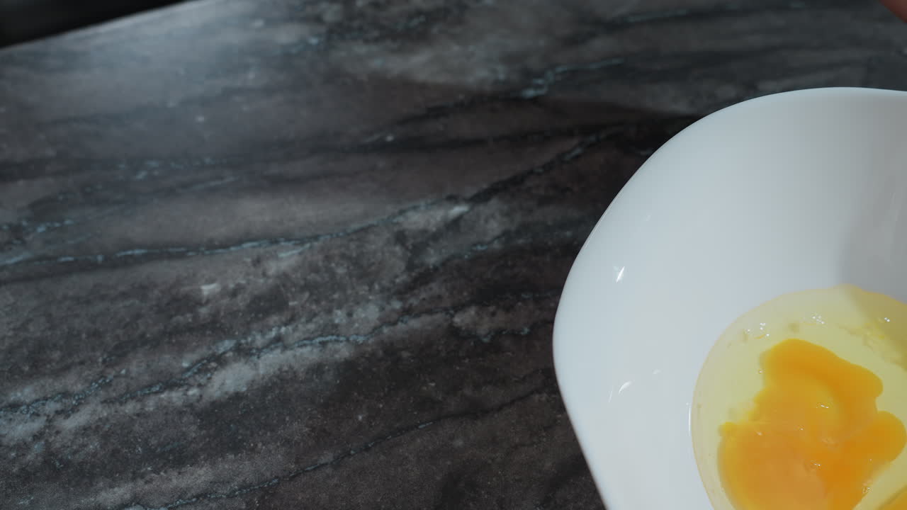 カウンタートップの上で卵を割る準備をしている人の手のクローズアップ近くに壊れた卵を含む白い鉢があり生卵黄と卵白が見えます