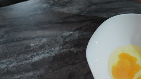 primer plano de la mano de una persona que se prepara para romper un huevo en una encimera, con un cuenco blanco que contiene huevos rotos cerca, la yema de huevo cruda y la blanca de huevos son visibles