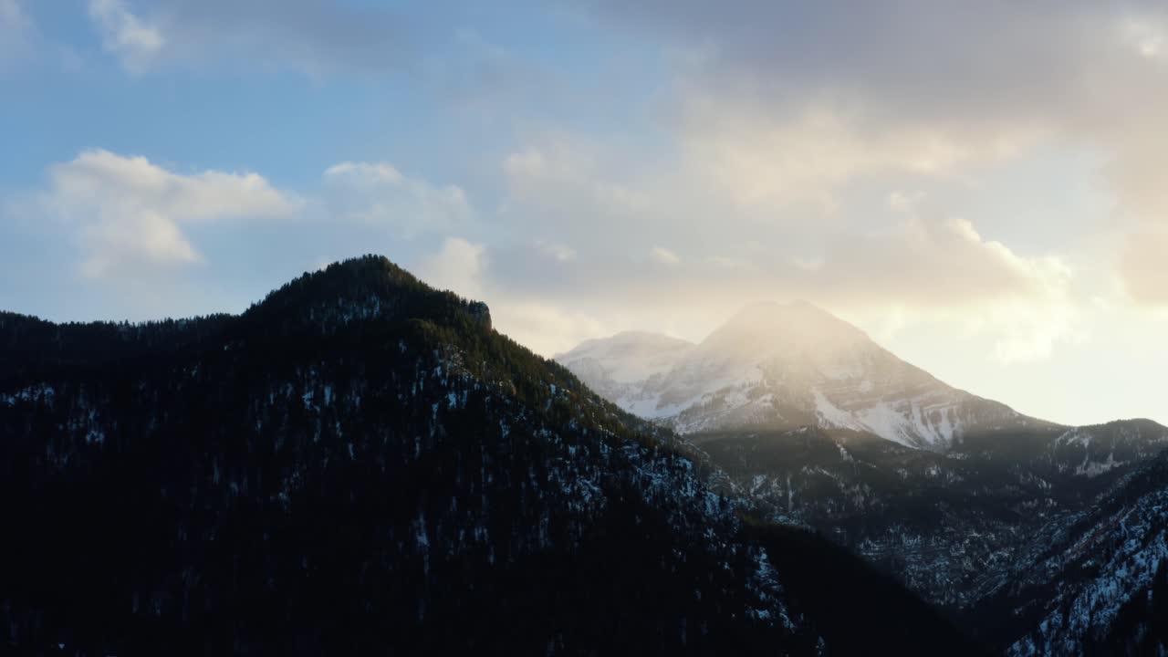 Dronekuva talvisesta maisemasta, jossa Timpanogos-vuori taustalla, mäntymetsän ympäröimänä auringonlaskun aikaan, jäätyneeltä Tibble Fork -tekojärveltä American Fork Canyonissa, Utahissa.