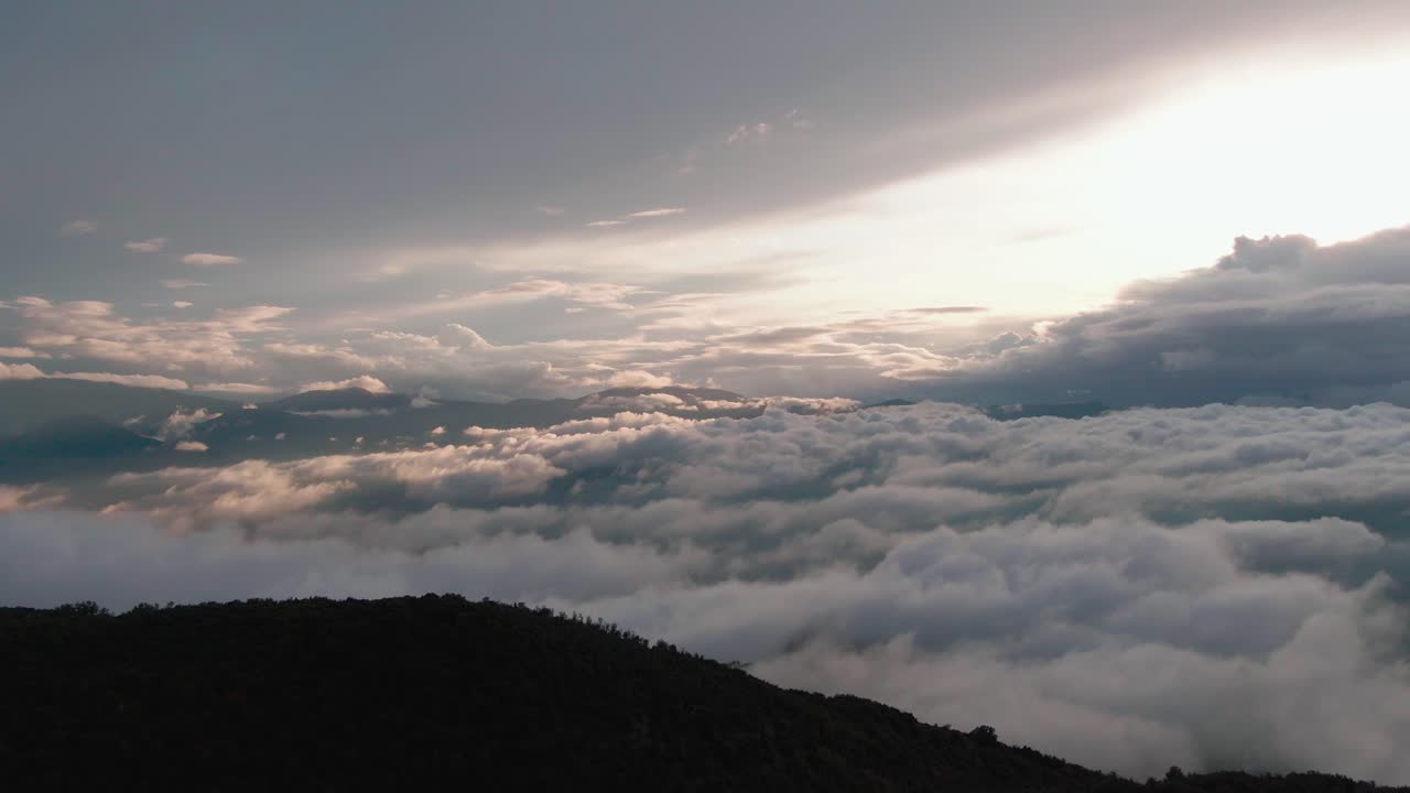 Overhead ascending flight above beautiful quaint mountain village on plateau with epic scenic views of cloudscape and bright white light from sun, rising aerial