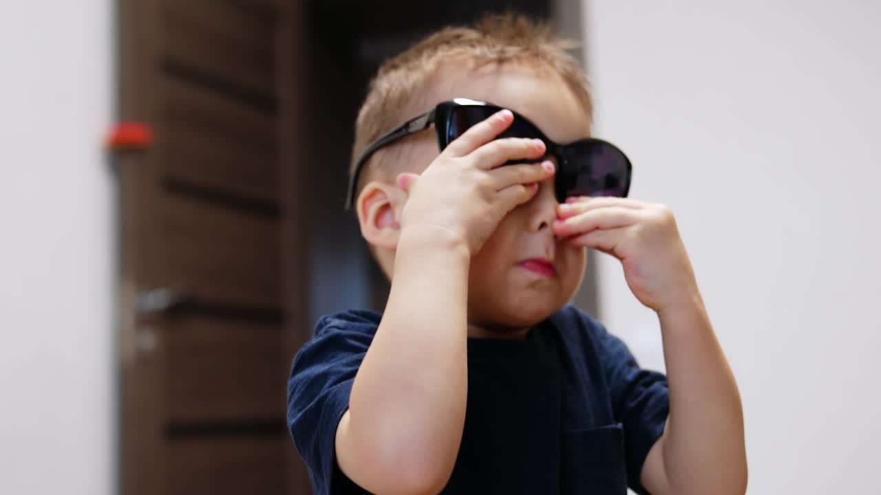 Cute Caucasian baby boy puts on sunglasses big for him. Close up portrait of a child at home.