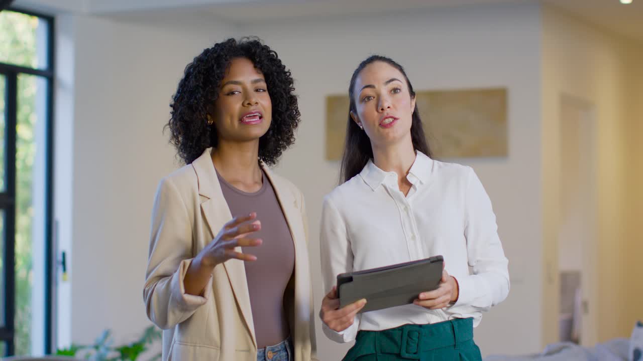 Diverse female coworkers reviewing plan at home after opening tablet, wearing beige blazer, scarf