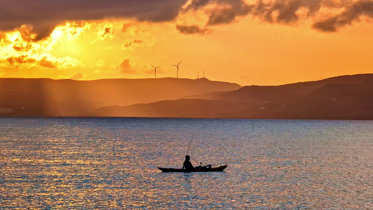 A lone fisherman watches a golden sunset from his small boat near the Sitia, Crete Greece with wind turbines on in silhouette on the mountains in the background