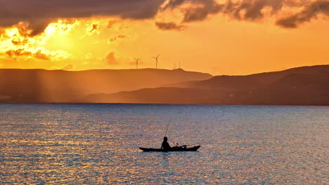 a lone fisherman watches a golden sunset from his small boat near the sitia, crete greece with wind turbines on in silhouette on the mountains in the background