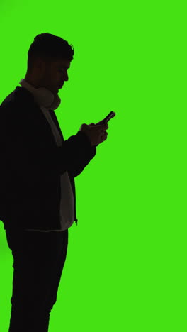 Vertical-Video-Silhouette-Of-Man-With-Wireless-Headphones-Text-Messaging-On-Mobile-Phone-Against-Green-Screen