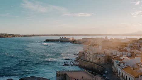 sunset colors with sunrays in the lens overlooking ortigia island, the old town of syracuse in sicily. historic buildings by the mediterranean sea with large waves crashing.