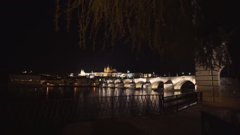 the prague castle and charles bridge over river vltava in the historical centre of prague,czechia,lit by lights at night,viewed from the other side of the river,pedestal shot from under a willow tree