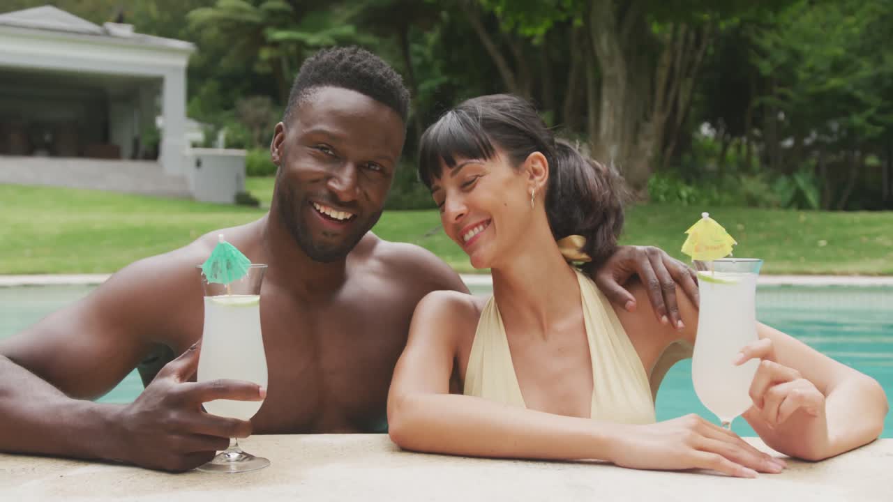 Ritratto di una coppia felice e multiculturale che indossa costumi da bagno e beve drink in piscina in giardino.
