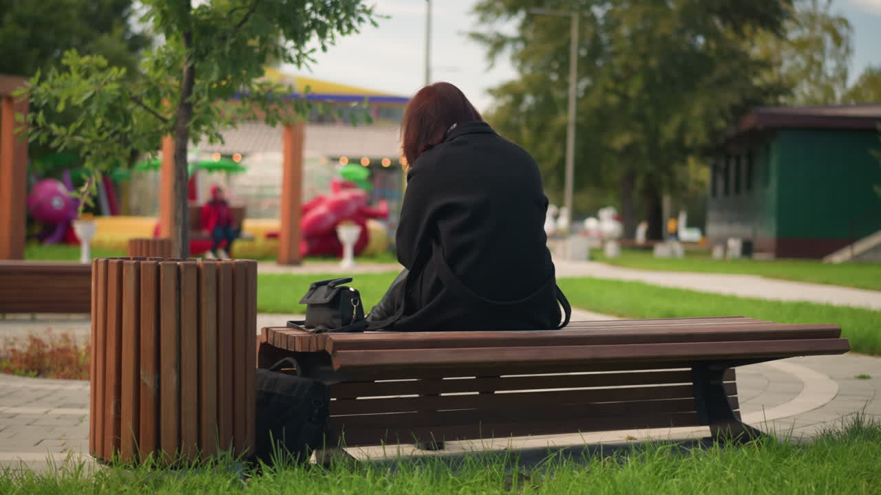 공원 벤치에 앉아있는 여성의 뒷면, 검은 코트와 부츠를 입고, 편안한 도시 공원 분위기에서 푸른 나무로 둘러싸여, 놀이터와 다른 공원 특징으로 흐릿한 배경