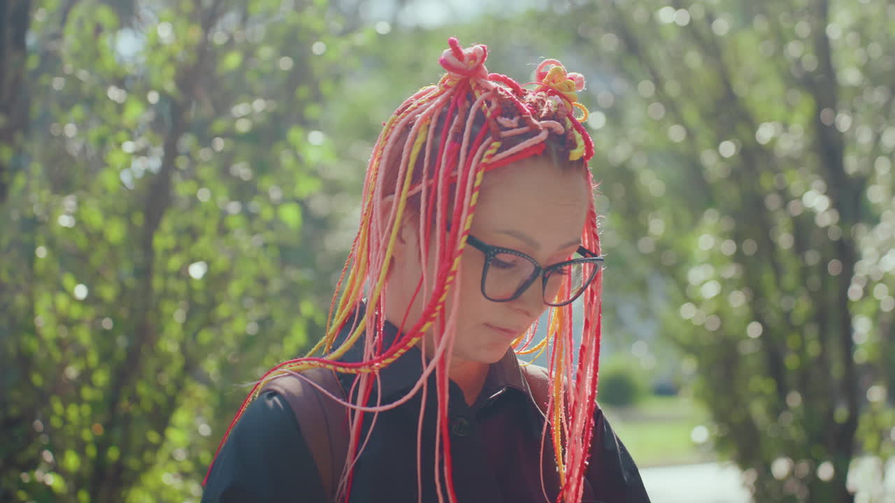 햇빛이 비치는 공원에서 핑크색 땋은 머리를 하고 아래를 내려다보는, 사색에 잠긴 백인 여성, 안경, 사려 깊은 창의적 에너지, 작가 또는 예술가의 생각을 비추는 클로즈업 인물 사진, 부드러운 보케와 잎이 무성한 배경