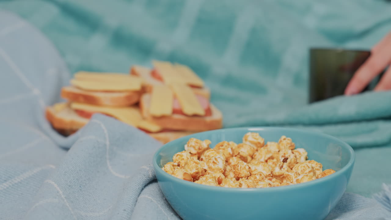 Close up of caramel popcorn in blue bowl placed on soft blanket with sliced bread sandwiches in background, relaxing indoor snack setting with cozy textures