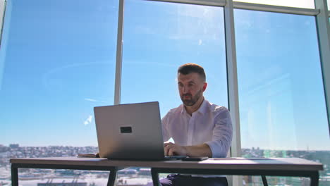 Handsome entrepreneur sitting at table in front of a laptop. Businessman working at his workplace in office on window backdrop.