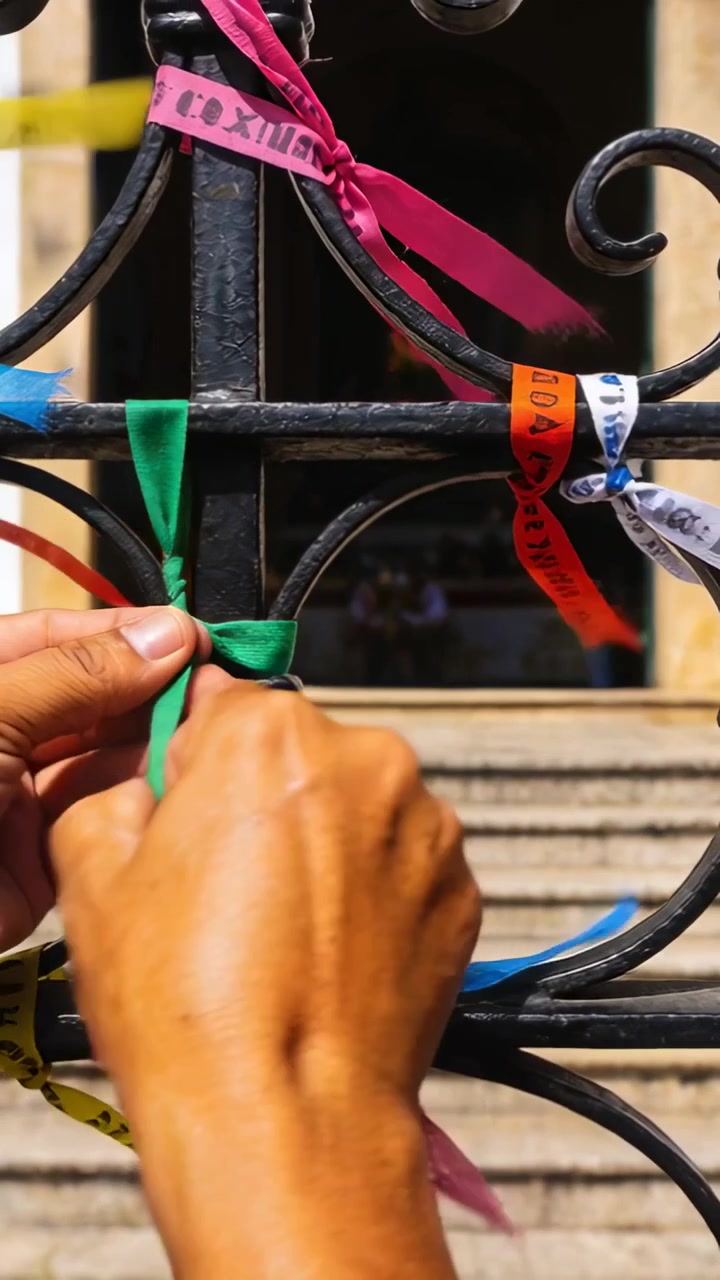 Video vertical: Brazos descubiertos sosteniendo una cinta verde y atando un nudo en una barandilla de hierro en una entrada, entre otras cintas.