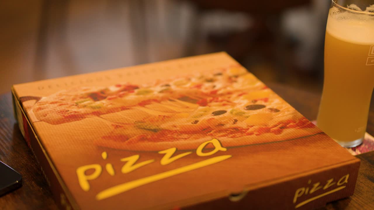 Hand opens takeaway pizza box revealing cheese, pepperoni, onion, green chili under warm indoor lighting