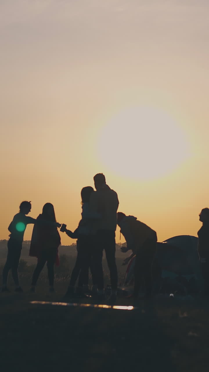 silhouettes of young people tourists spending time by tent against clear evening sky at orange sunset on horizon