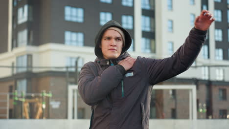 athlete turning head and stretching arm by placing right hand on left arm in preparation for sports activity, dressed in hoodie, standing confidently in urban sports arena