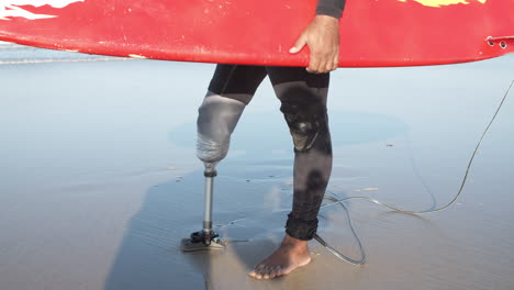 primer plano de un surfista irreconocible con una pierna artificial parada en la orilla del océano y sosteniendo una tabla de surf bajo el brazo