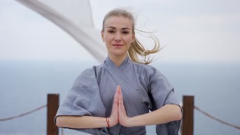 Portrait-of-smiling-female-coach-yoga-in-robe-standing-at-terrace-folded-hands-in-Namaste-gesture