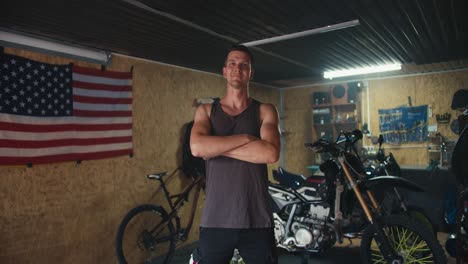 Portrait:-A-male-mechanic-in-a-gray-T-shirt-confidently-folds-his-arms-across-his-chest-in-his-workshop,-in-which-a-US-flag-hangs-on-the-wall-and-motorcycles-stand