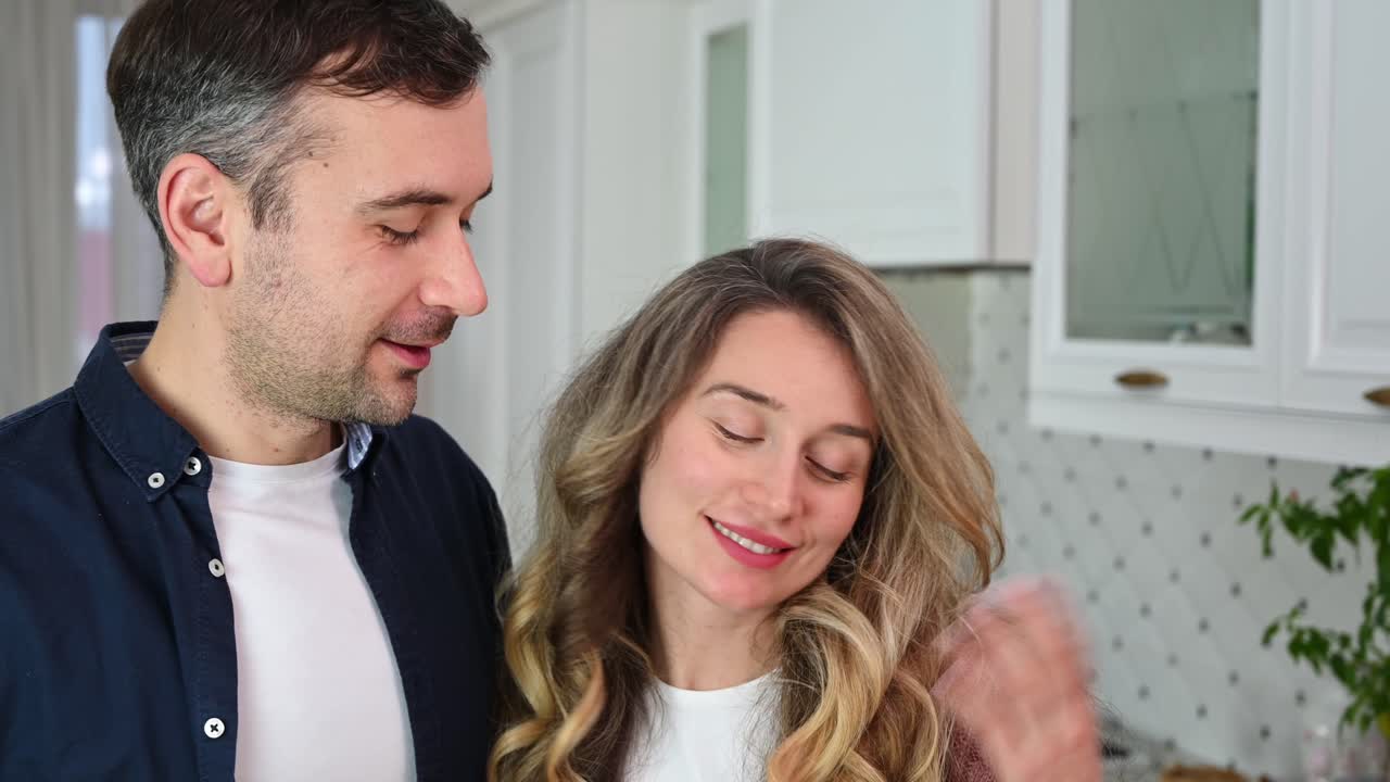 Una coppia matura affettuosa che parla e sorride in una moderna cucina bianca. Un uomo carismatico e una bella donna bionda si guardano con affetto. Romantico San Valentino. Concetto di relazioni e sentimenti