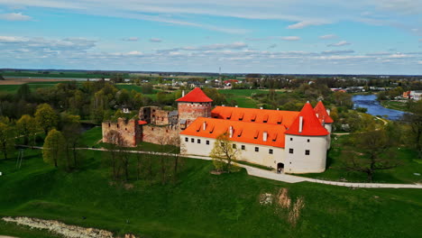 Aerial-drone-rotating-shot-over-historic-Bauska-Castle-Museum-in-Latvia-surrounded-by-lush-green-vegetation-during-evening-time