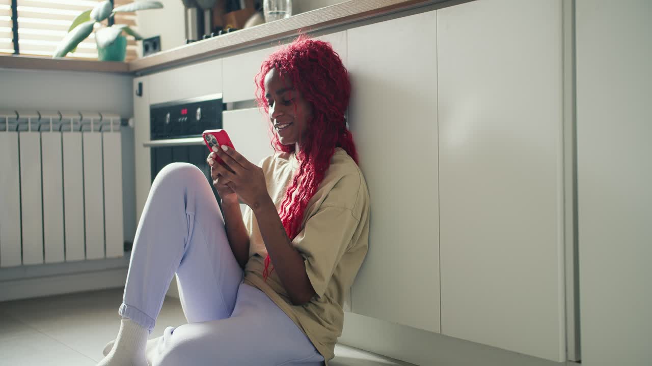 Joyful African American girl with red curly hair sitting on kitchen floor and srfing internet using mobile