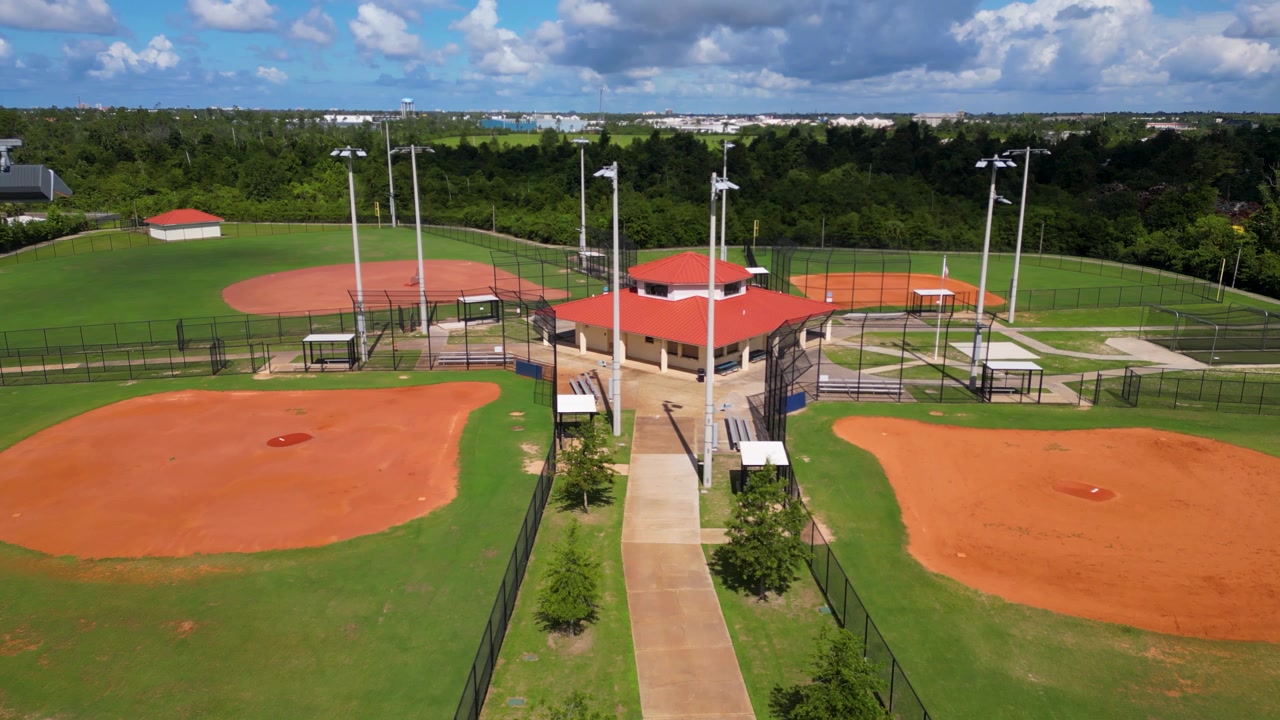 Ripresa lenta con drone che avanza lungo il marciapiede verso il chiosco, tra pali della luce e campi da baseball, in un piccolo parco.