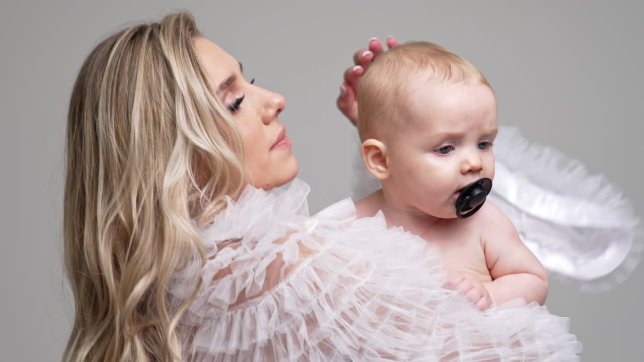 Loving mother in white robe waving her little child. Cute baby with pacifier in mouth starts to cry. White backdrop.