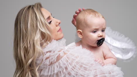 loving mother in white robe waving her little child. cute baby with pacifier in mouth starts to cry. white backdrop.