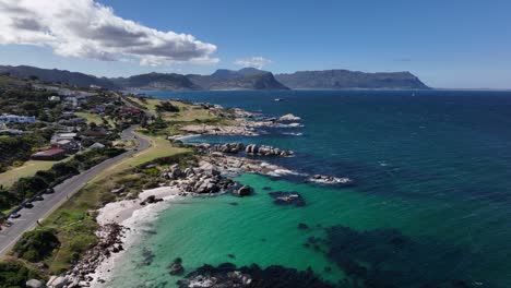 Wunderschöne-Luftaufnahme-Einer-Straße-Entlang-Der-Felsigen-Küste-Mit-Tiefgrünem-Wasser,-Häusern,-Bergen-Und-Klippen-Im-Hintergrund-An-Einem-Sonnigen-Tag-In-Der-Nähe-Von-Kapstadt,-Südafrika