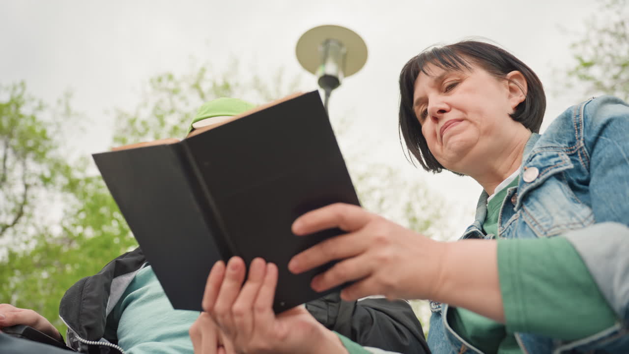 Adult And Teenager Reading Together Outdoors On Bench With LowAngle View Book Held Between Them, Attentive Expressions, Denim Jacket And Cap Visible, Lamppost Above, Shared Concentration