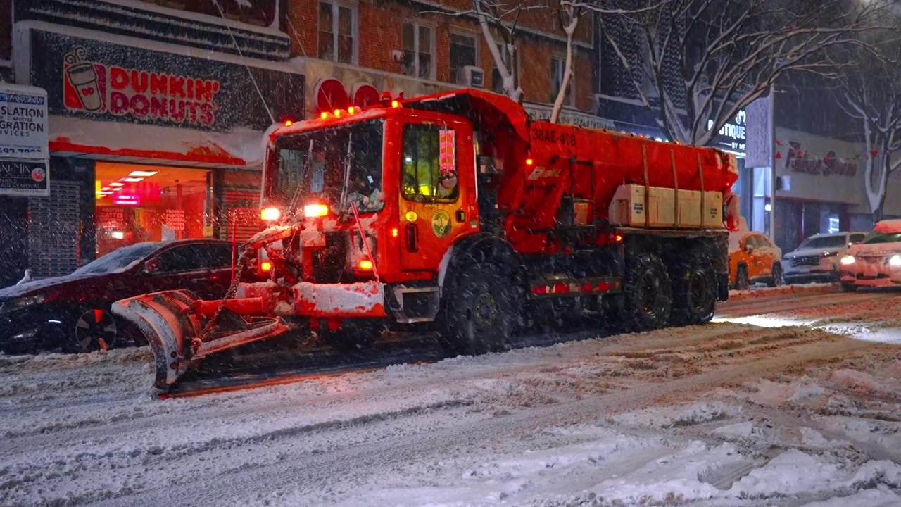 Nova York, EUA, 20 de fevereiro de 2026: Caminhão espalhador de sal vermelho limpando neve em rua urbana. Caminhão limpa-neve pesado estacionado em frente a uma cafeteria durante uma nevasca noturna em Nova York