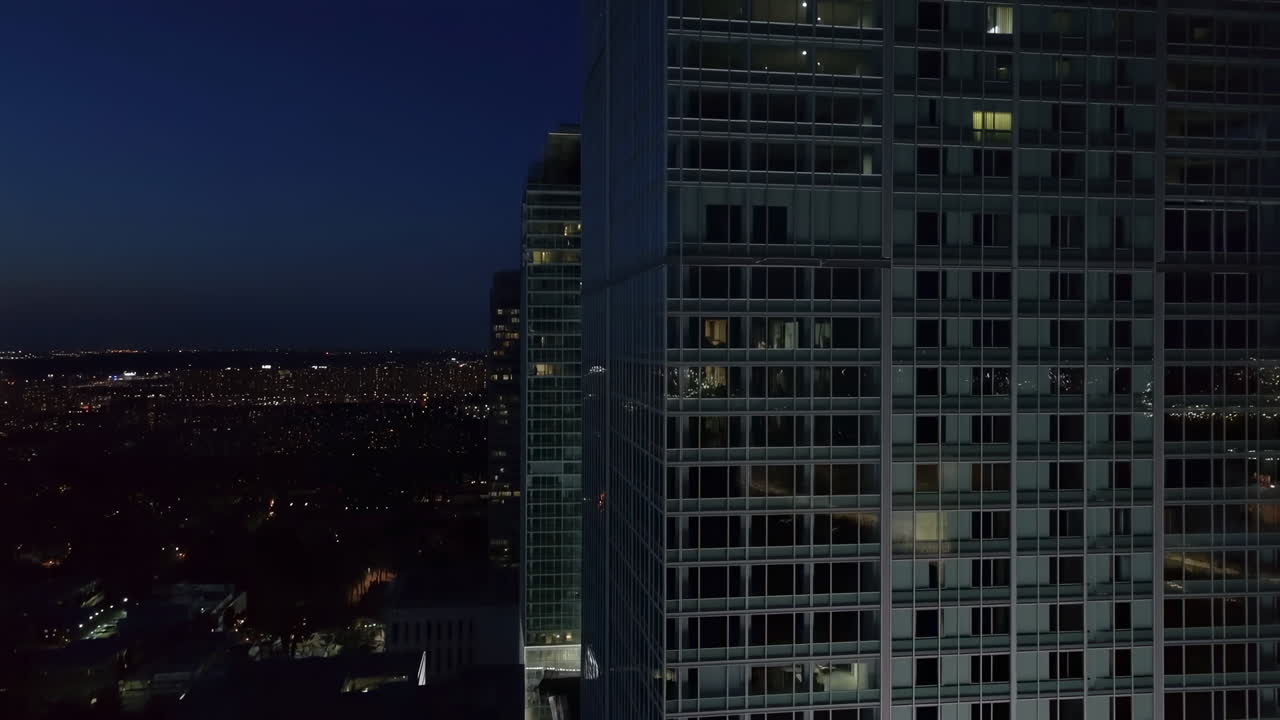 Paisagem urbana noturna com janelas de arranha-céus iluminando o horizonte, drone aéreo revela a fachada de vidro iluminada de um edifício moderno.